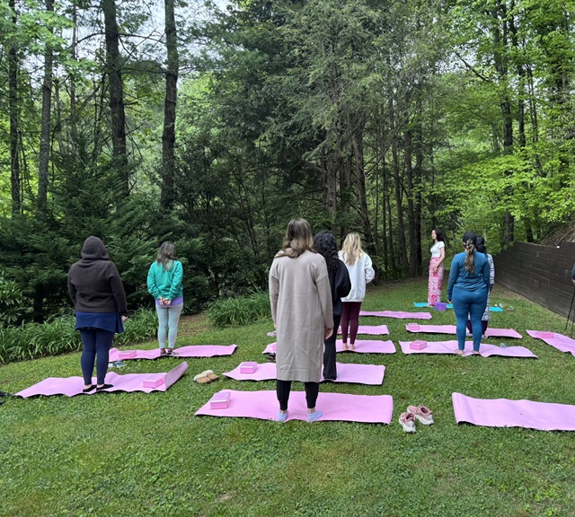 Women standing in a semicircle on pink mats among tall trees during an outdoor practice